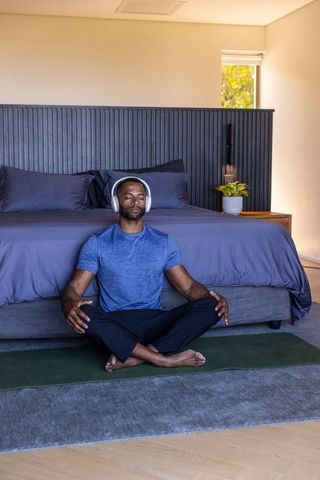 Mature african man meditating cross-legged with headphones on yoga mat in modern bedroom