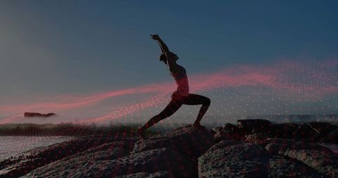 Silhouette woman practicing warrior lunge on rocky shore at dusk with red digital overlay