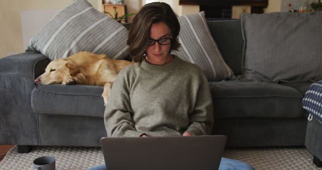 Young woman sitting on the floor and working on a laptop in a cozy living room setting while a dog sleeps on the sofa behind her. Ideal for illustrating concepts of remote work, home lifestyle, pet-friendly homes, technological connectivity, and leisure time at home. Great for websites or articles about working from home, comfortable home design, balancing work and life, and pet companionship.