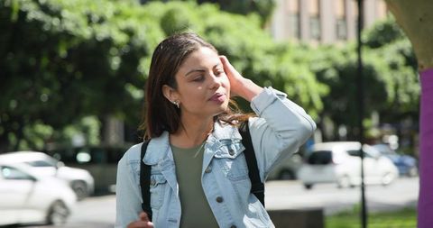 Woman adjusting hair on city sidewalk wearing denim jacket and backpack