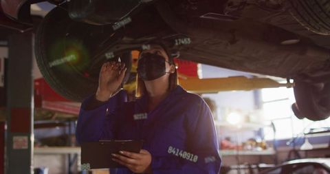 Female mechanic inspecting car undercarriage on lift holding clipboard and flashlight, masked