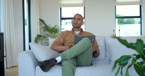 Man relaxing on modern gray sofa holding tablet in bright minimalist living room