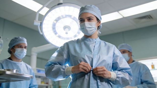 Female surgeon securing sterile gown in operating room under surgical light with team