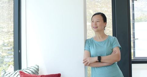 Asian woman leaning by window in modern bright living room wearing teal tee and smartwatch