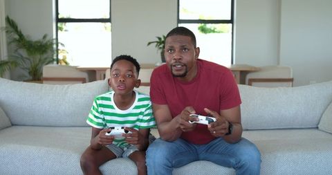 African american father and son playing video game on sofa in modern living room
