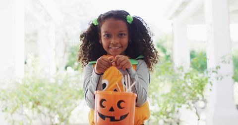 Smiling girl holding pumpkin bucket on porch wearing pumpkin costume for halloween