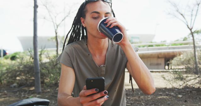 Fashionable biracial man with dreadlocks using smartphone and drinking coffee. Street style, modern urban lifestyle and communication.