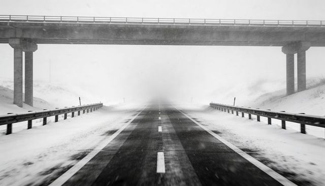 Desolate two-lane highway vanishing into whiteout beneath concrete overpass, monochrome storm