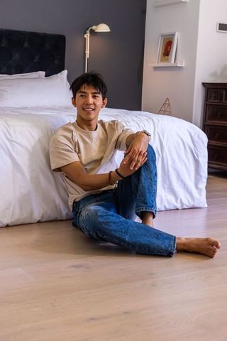 Smiling asian man sitting on bedroom floor with knee drawn up wearing wristwatch