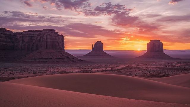 Sunrise casting lavender light over sand dunes and three sandstone buttes