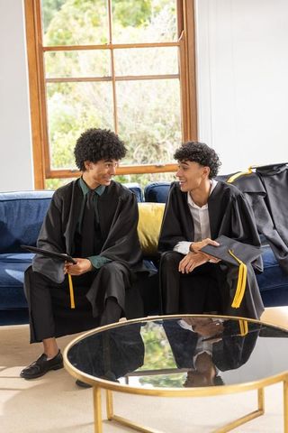 African american graduates sitting on sofa in robes sharing joy and mortarboards
