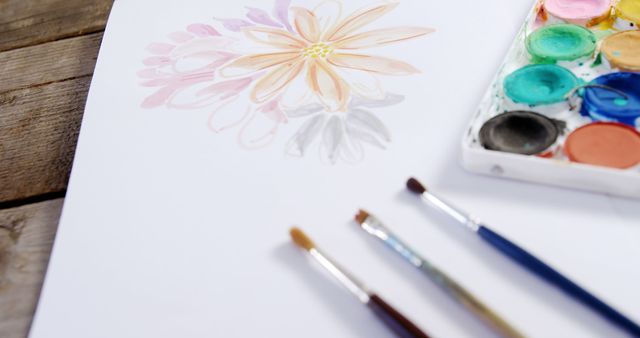 A watercolor painting of a flower is in progress beside a palette with vibrant colors, with copy space. Art supplies including brushes suggest a creative session, by an artist or hobbyist.