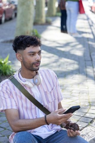 Sunlit asian man sitting on curb using smartphone and headphones while enjoying pastry