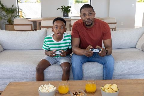 African american father and son playing video games on sofa, sharing snacks and bonding