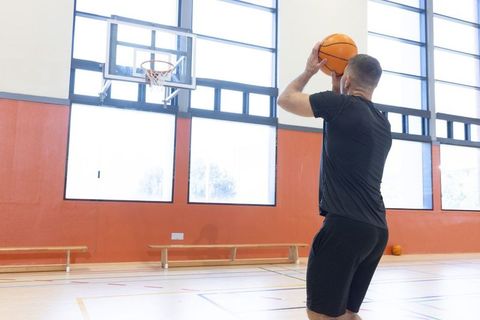 Male athlete shooting basketball in bright indoor gym with high windows and wall-mounted hoop