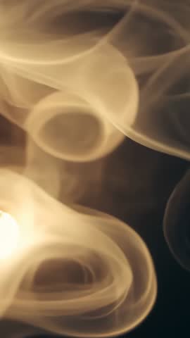 Swirling golden smoke rings dancing around backlit lamp in dark studio vertical close-up video