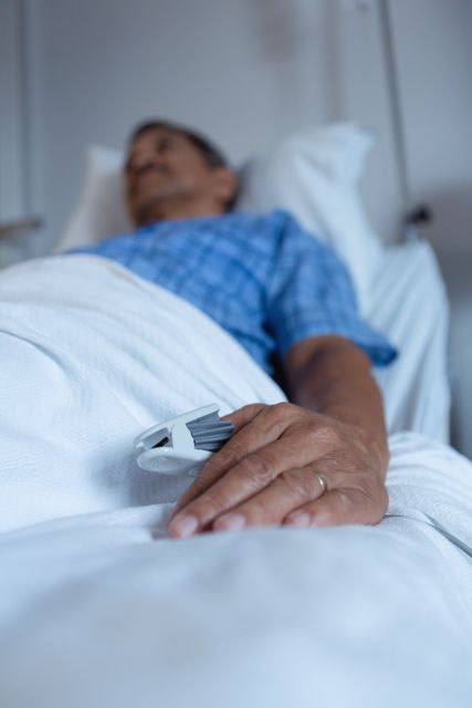 Mature male patient lying in a hospital bed receiving treatment. He is wearing a hospital gown and has a medical device attached to his finger for monitoring. This image can be used for healthcare, medical care, patient recovery, and hospital-related content.