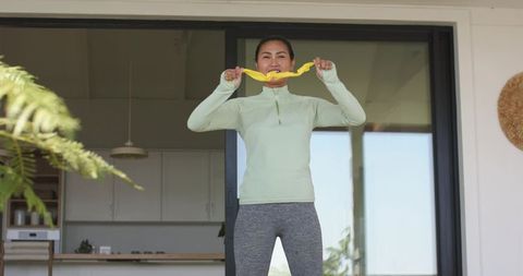 Asian woman stretching with yellow resistance band in sunlit home doorway