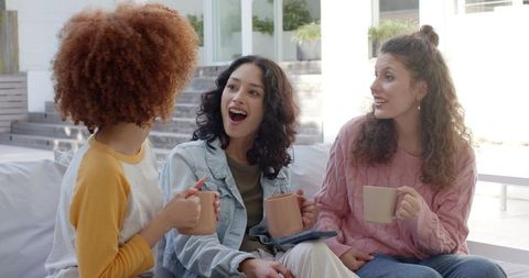 Friends chatting on sunny patio sofa holding mugs and tablet casual weekend gathering