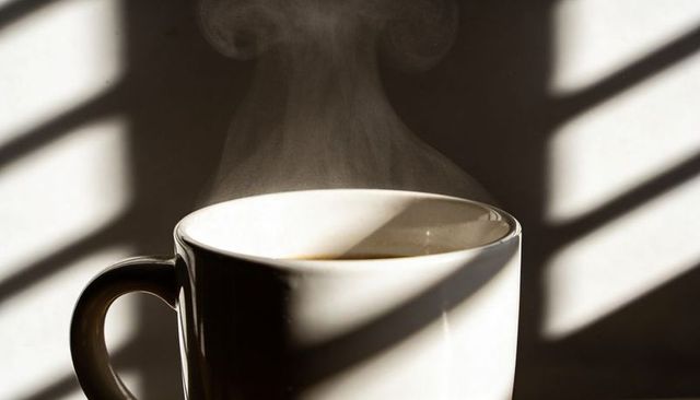 Steaming white ceramic coffee mug with dramatic window blind shadows morning light