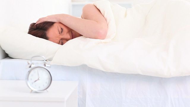 A woman is covering her ears, struggling to wake up with an alarm clock by her bedside, a scene commonly faced during early mornings. This visual can be utilized for concepts related to difficult mornings, insomnia, need for relaxation, or promoting morning aids.