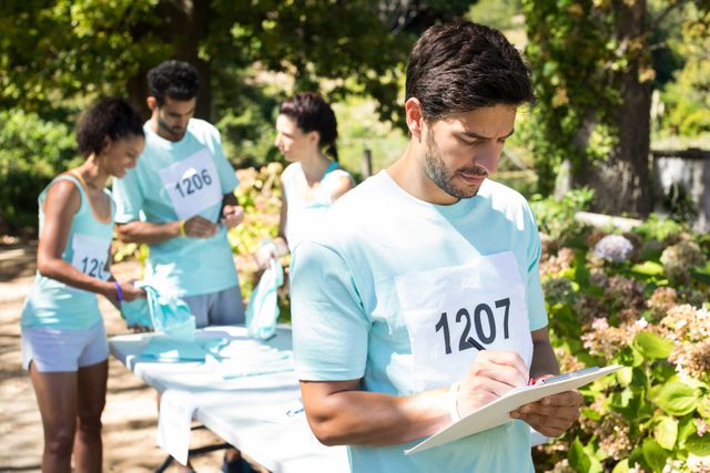 Participants are registering for a marathon at a check-in table in a park. They are wearing numbered bibs and filling out papers. The scene suggests community engagement, active lifestyle, and organized sports events. Use for promoting fitness events, athletic competitions, and community sports activities.