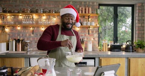 Mid-adult african american man whisking in bowl wearing santa hat in cozy rustic kitchen