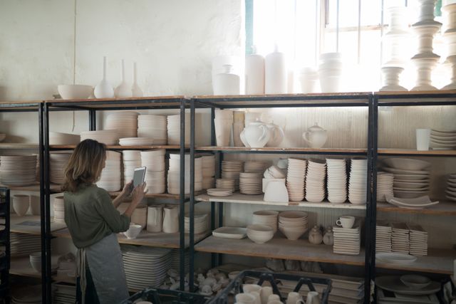 Image depicts female potter using digital tablet while standing near shelves filled with pottery in workshop. Highlights modern technology in traditional craft setting. Suitable for illustrating themes of creative processes, ceramics, artisans, and technology in the arts. Can be used in blog posts about pottery-making, small business operations, and artisanship.