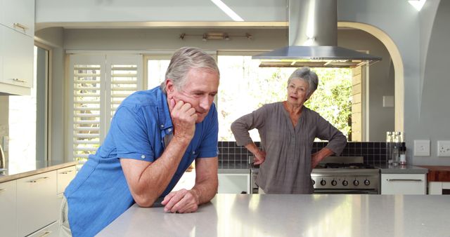 Elderly couple having an argument in a modern kitchen with island counter. The man looks down with a frustrated expression, while the woman stands in the background with her hand on her hip. This image can be used to represent conflict in relationships, challenges in long-term relationships, family disputes, or senior living dynamics.