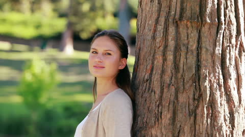 Smiling Young Woman Leaning on Tree in Sunlit Park