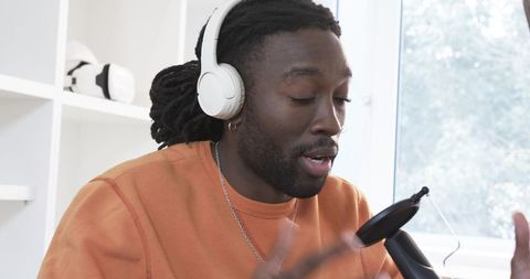 African american man recording podcast at home studio wearing white headphones and speaking