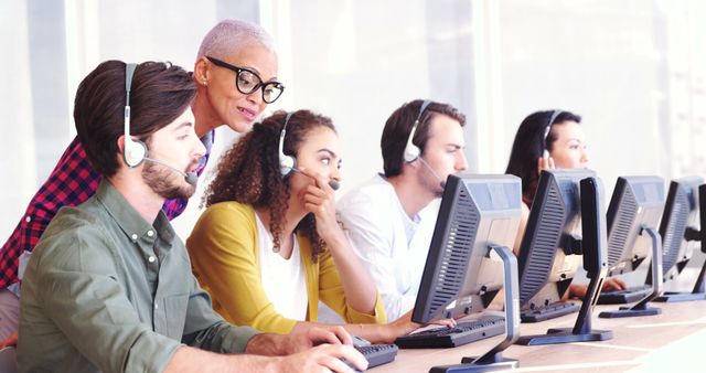 A diverse group of customer service representatives, including Caucasian, African American, and Latino individuals, are working at a call center, with copy space. They are equipped with headsets and focused on their computer screens, providing support to clients.