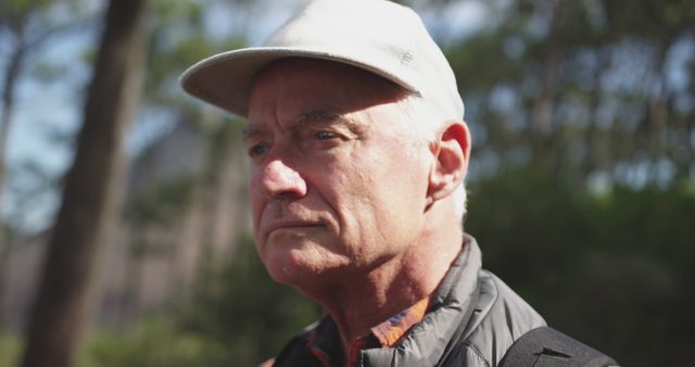 An elderly man wearing a white cap stands in an outdoor setting, displaying a serious and reflective facial expression. The background features blurred greenery, indicating a forest environment. This image is suitable for use in articles or advertisements focused on senior life, nature exploration, outdoor activities, or personal narratives involving mature individuals.