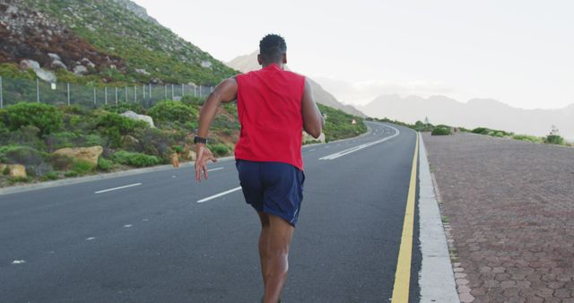 Image shows a man running on an empty mountain road. He is wearing a red tank top and blue shorts. There is a lush green landscape and mountains in the background, making the scenery picturesque. This image can be used for promoting fitness, outdoor activities, and training programs. It can also be used in articles or blogs about healthy lifestyle, motivation, and exercise routines.