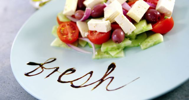 A plate of Greek salad is garnished with the word fresh written in balsamic reduction, with copy space. The vibrant colors of the tomatoes, cucumber, feta cheese, and olives emphasize the freshness of the ingredients.