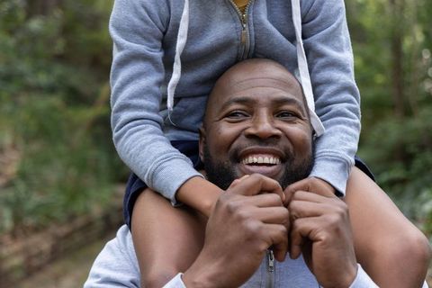 African american dad carrying child on shoulders laughing together on forest trail