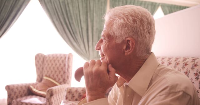 Elderly man with white hair is relaxing and smiling, sitting comfortably at home in soft light. He is deep in thought, giving an impression of wisdom and tranquility. This stock can be used for themes related to aging, retirement, senior healthcare, peaceful living, or promoting home comfort.