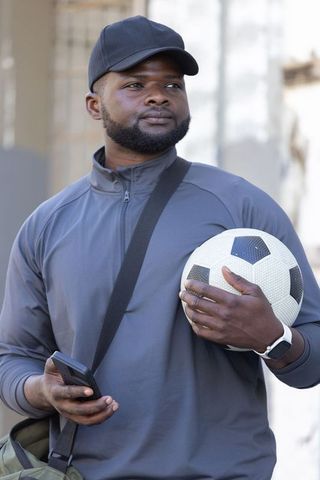 Street athlete holding soccer ball and checking smartphone wearing cap and gray jacket