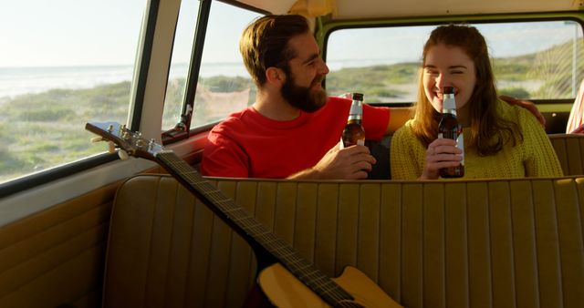 Young couple sitting inside camper van, enjoying beers and laughing during sunset. Picturesque landscape of countryside visible through van windows. Perfect for lifestyle, travel, and adventure themed content. Ideal for promoting road trips, vacations, and products related to outdoor leisure and beverage consumption.