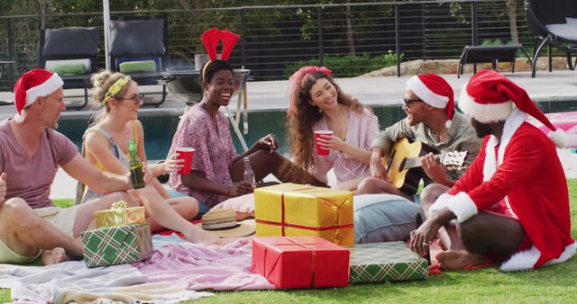 Multiracial group of friends celebrating Christmas outdoors. All wearing Santa hats and festive attire, sitting on a blanket with presents. One is playing a guitar, while others are laughing and holding drinks. Perfect for ads or content focusing on festive celebrations, multicultural gatherings, outdoor holidays, and friendship.