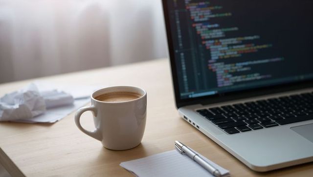 Steaming coffee mug with crema beside laptop showing code and notepad on light wood desk