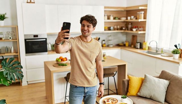 Young man taking selfie in bright modern kitchen smiling in striped tee casual lifestyle