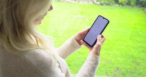 Rearview Woman Holding Smartphone in Garden, Wearing Cream Sweater, Green Lawn Background