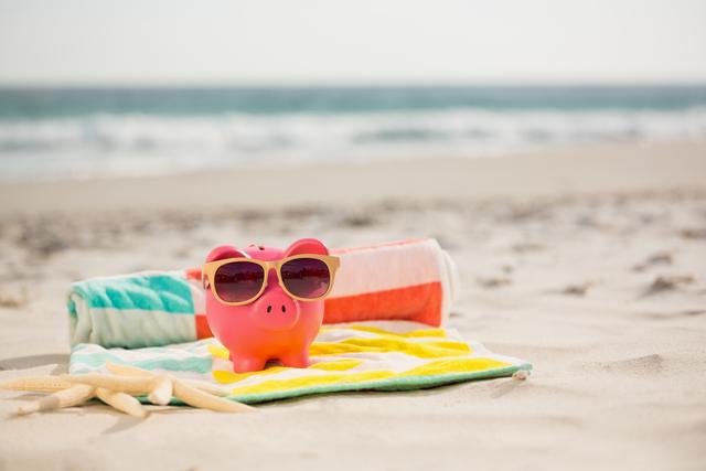 Piggy bank wearing sunglasses on colorful beach blanket with starfish on sandy beach. Ideal for concepts related to summer savings, vacation planning, financial planning, and relaxation. Perfect for travel agencies, financial institutions, and summer promotions.