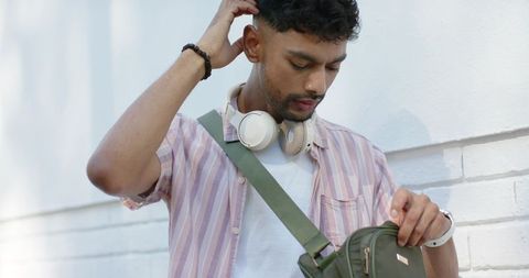 Urban Asian Man Wearing Headphones and Crossbody Bag Opening Backpack by White Brick Wall