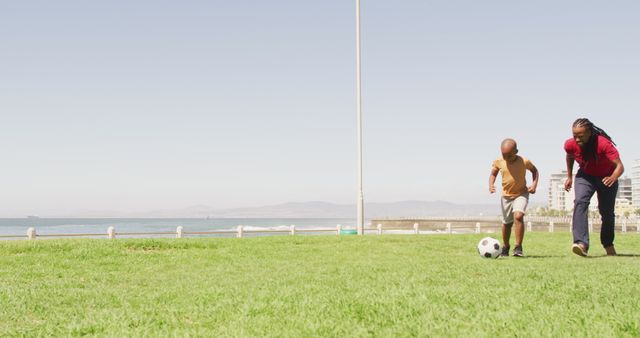 Brown-skinned father and young son playing soccer on grassy field beside the sea with bright blue sky in background. Perfect for illustrating family bonding, outdoor activities, healthy lifestyle, parent-child relationship, summer fun and fitness themes.