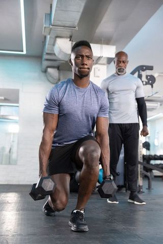African american trainer coaching young man performing weighted dumbbell lunges in gym