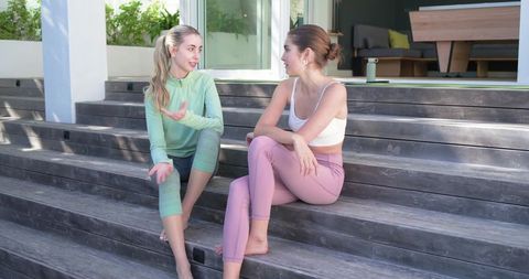 Young women in activewear sitting on outdoor wooden steps talking after workout, lifestyle