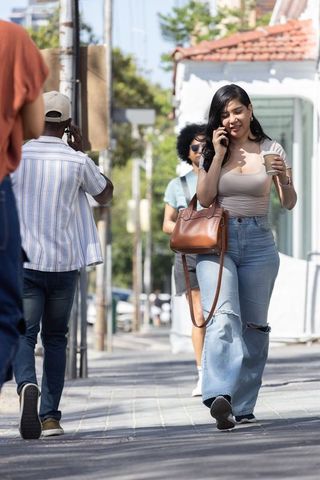 Hispanic woman walking downtown talking on phone while holding coffee and leather bag