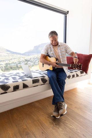 Senior man tuning acoustic guitar on window bench with laptop and panoramic mountain view